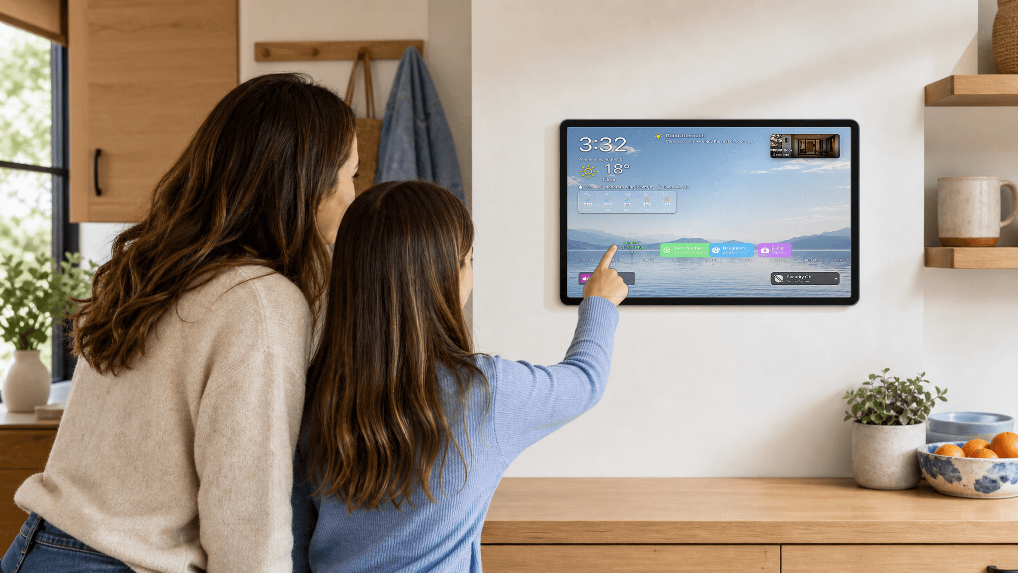A mother and daughter checking their family calendar on a wall-mounted iPad in a sunny kitchen